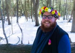 Man with a beard and glasses smiles at the camera outside while wearing a flower crown