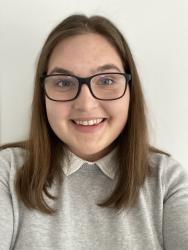 Headshot of Emily Mowat; woman with brown hair and glasses smiles at the camera wearing a grey sweater