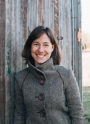 Woman with short brown hair waring a grey fall jacket leans against a wooden structure, smiling