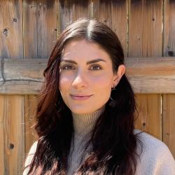 Justina has long dark hair. She is wearing a grey turtleneck, standing in front of a wooden fence, smiling at the camera.