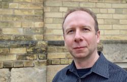 A man with short hair wearing a blue collared shirt in front of a yellow brick wall