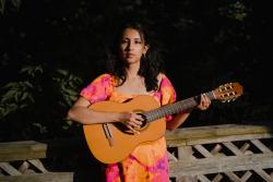 Womas with dark hair wearing a pink and orange dress holds a guitar