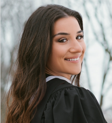 A woman with dark hair wearing a graduation gown looks over her shoulder and smiles