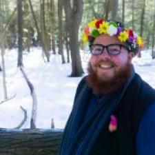 Man with a beard and glasses smiles at the camera outside while wearing a flower crown