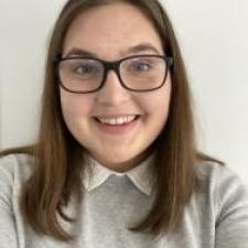 Headshot of Emily Mowat; woman with brown hair and glasses smiles at the camera wearing a grey sweater