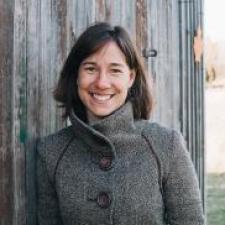 Woman with short brown hair waring a grey fall jacket leans against a wooden structure, smiling