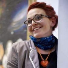 Jemma has red hair and wears glasses, a blue scarf, and a red heart necklace. She is smiling, looking just beyond the camera.