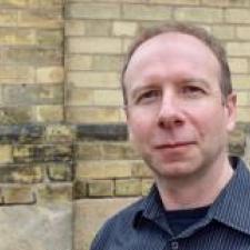 A man with short hair wearing a blue collared shirt in front of a yellow brick wall