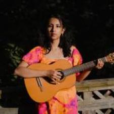 Womas with dark hair wearing a pink and orange dress holds a guitar