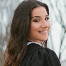 A woman with dark hair wearing a graduation gown looks over her shoulder and smiles