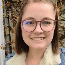 Individual with short brown hair and glasses wearing a denim glasses, smiling