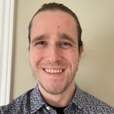 A man with his hair pulled back wearing a blue patterned button up, smiling