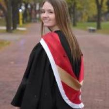 Woman with brown hair wearing a black graduation gown and red and gold sash looks over her shoulder and smiles