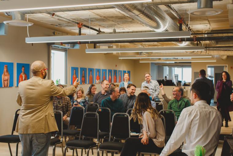 A bald man gestures at a smiling audience in a medium-sized room