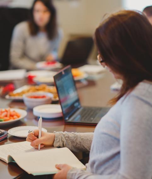 Brown-haired woman writes in a notebook at a large table, surrounded by others