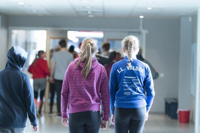 Two people walk down a hallway, visible from the back. One is wearing a purple top, one is wearing a blue top.