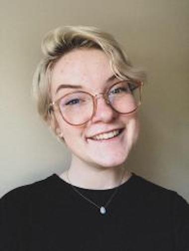 Person with short blonde hair, large glasses and black shirt smiles at the camera.