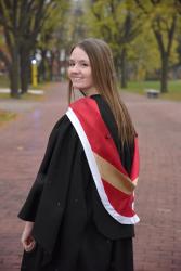 Woman with brown hair wearing a black graduation gown and red and gold sash looks over her shoulder and smiles